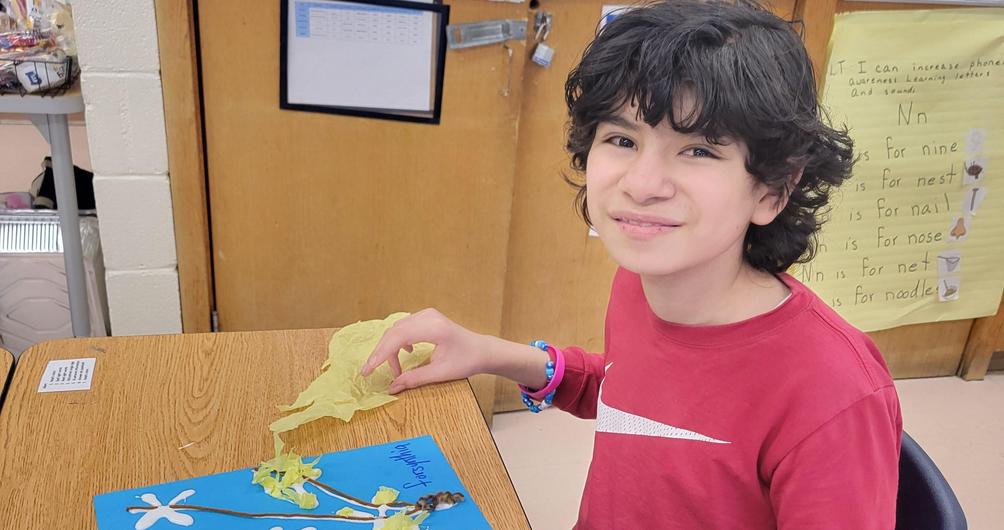 Boy creating art with yellow tissue paper and sticks on blue poster board.