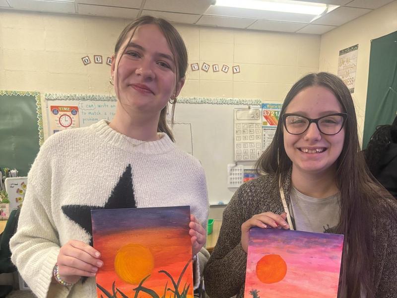 Two girls holding paintings of sunset scenes.