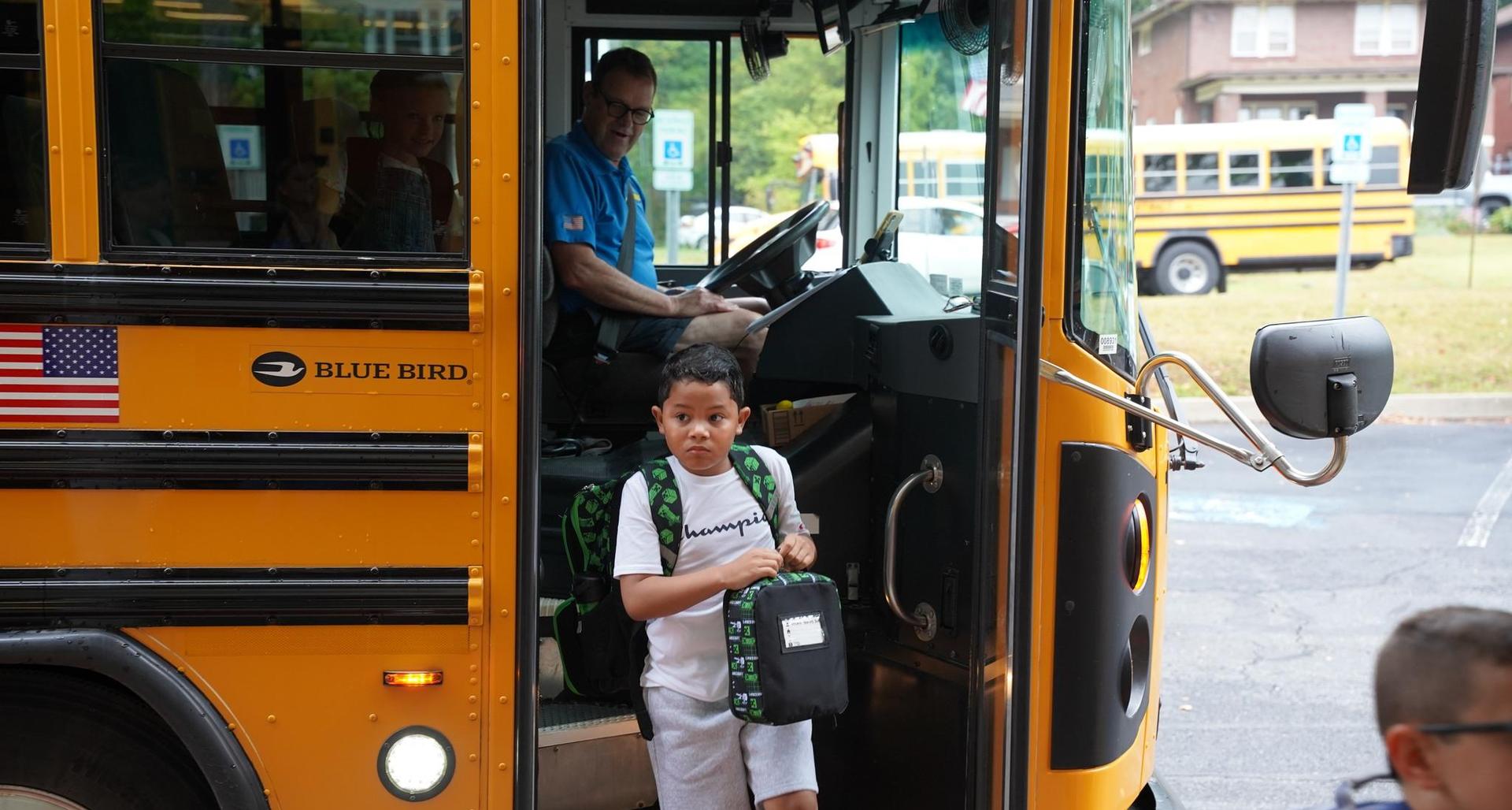 A boy with a backpack exits a school bus while the driver sits inside.