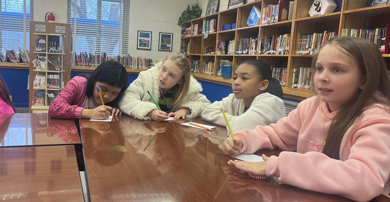 Five children sitting at a table in a library, engaged in writing or drawing.