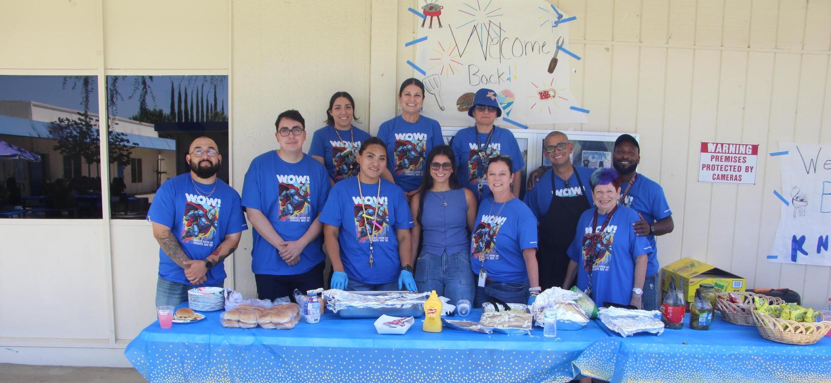 Group of people in blue shirts gathered around a table with food and drinks.