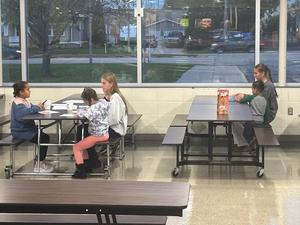 Four children sit at tables in a school cafeteria, engaged in various activities.