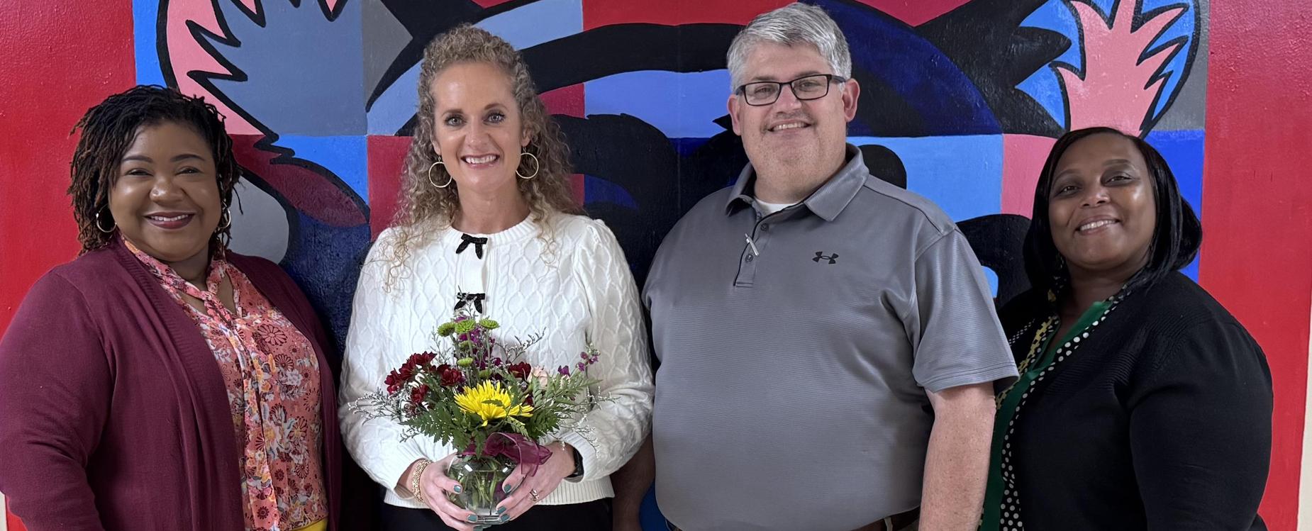 Smiling group of four people with a flower vase in front of a bright, artistic background.