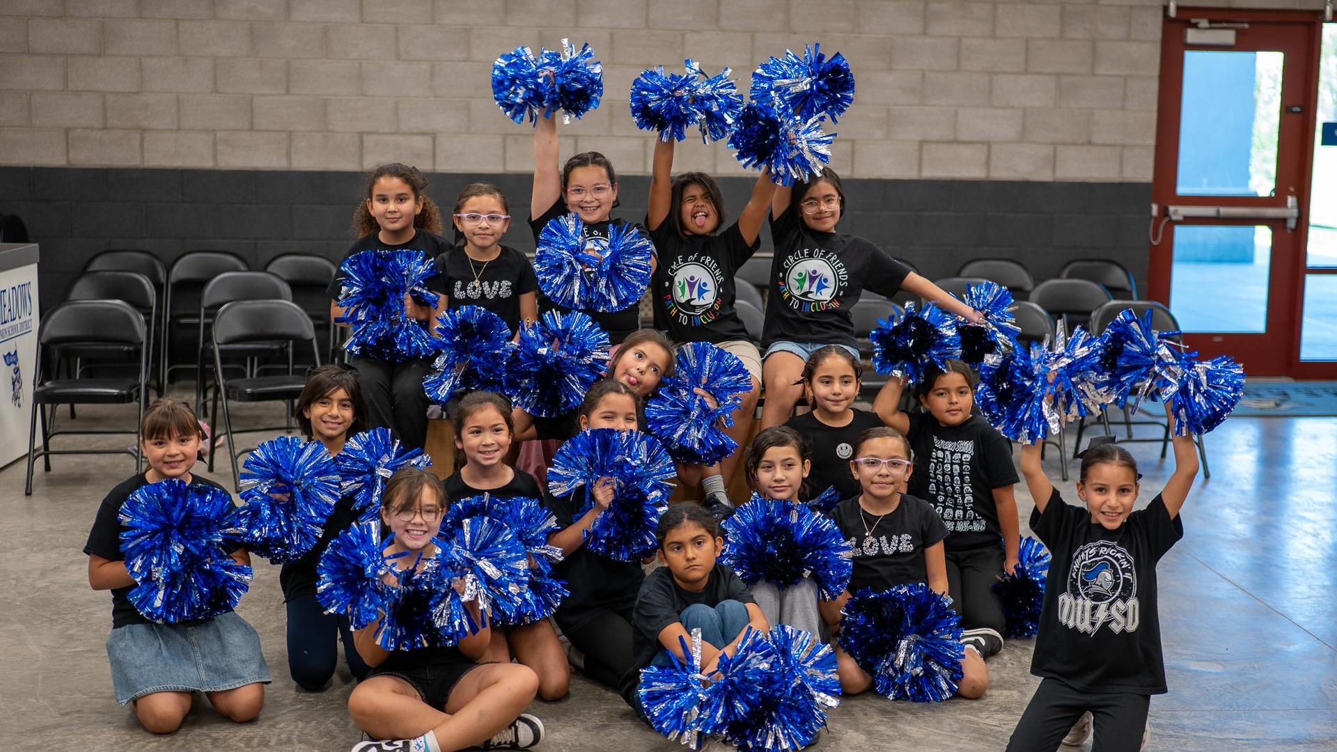 A group of girls in black shirts cheer with blue pom-poms, posing joyfully together.