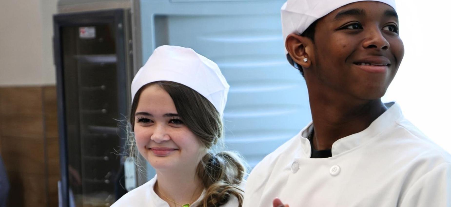 Two young chefs wearing uniforms and smiling in a kitchen environment.