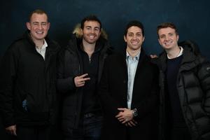 Four men posing together with smiles and gestures, in warm jackets against a blue backdrop.
