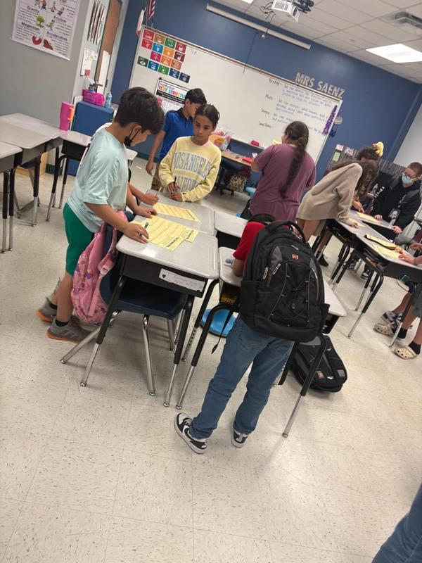 Boys and girls standing around classroom desks writing on yellow papers