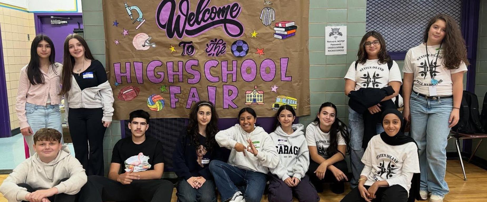 students sitting on floor and standing by sign for high school fair
