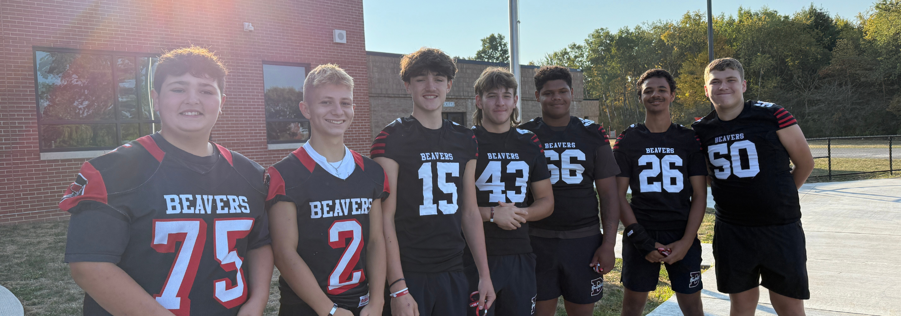 A group of seven young football players in black uniforms standing together outside on a sunny day.