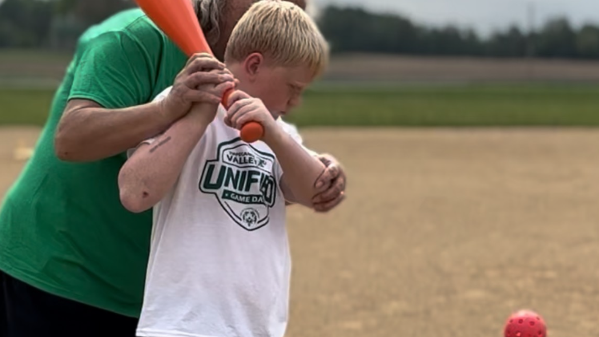 Child learning to bat with assistance from an adult on a sports field.