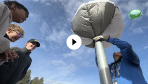 Middle Schoolers watch as a teacher inflates a small hot air balloon on a metal pipe.