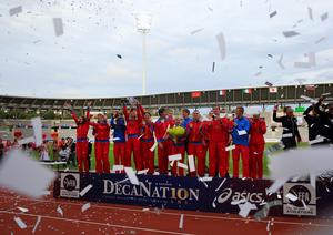 Athletes in red uniforms celebrating with trophies on a podium, confetti falling around them.