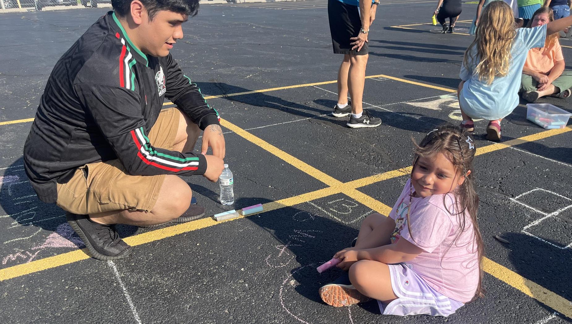 A young girl enjoys chalk drawing on asphalt, assisted by an older boy.
