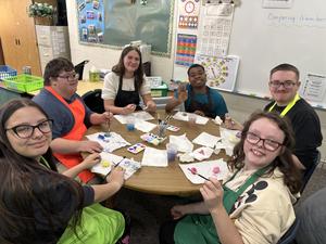 A group of six students painting at a table, each using different colors and materials.