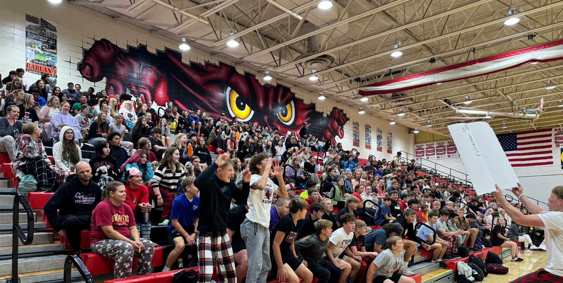 Crowd of students cheering in a gym with a large mural of a dragon on the wall.