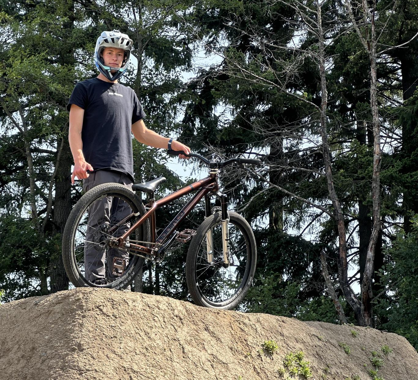 Young person wearing a helmet stands on a bike on a dirt mound surrounded by trees.