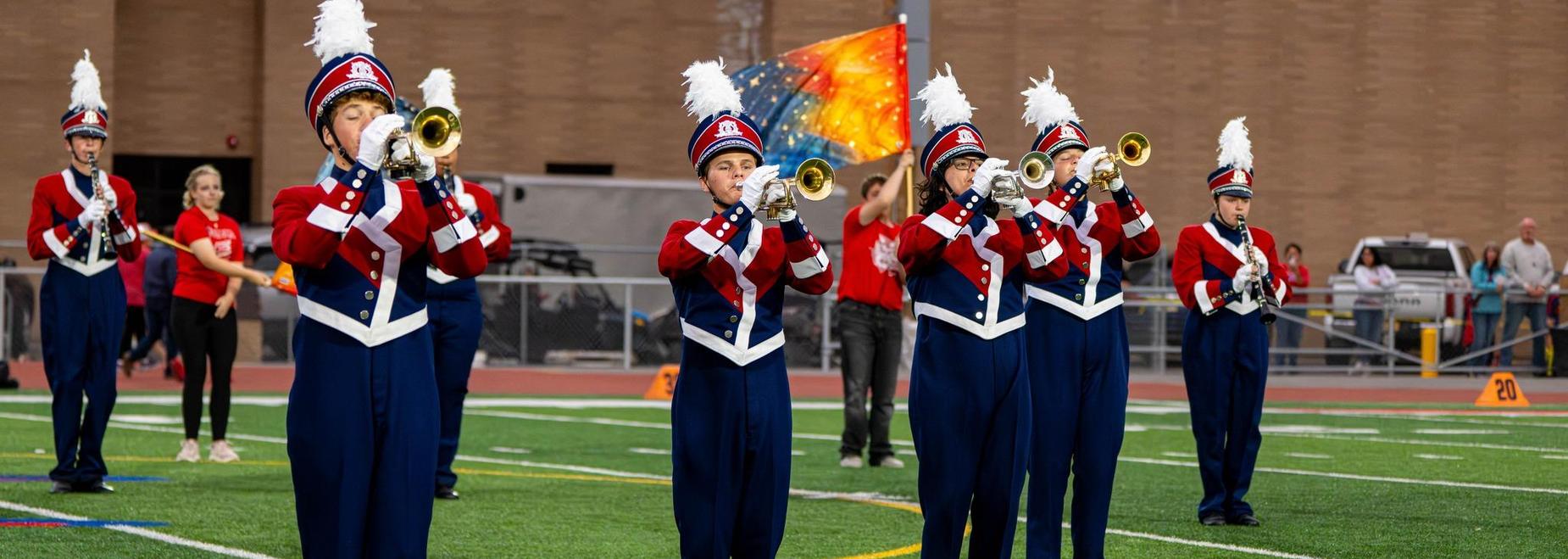 Marching band members in uniforms perform with trumpets on a sports field.