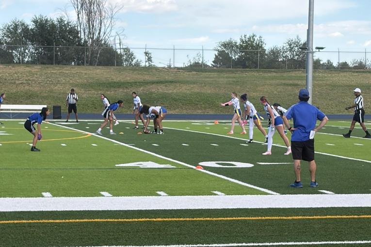 FLHS Players in a flag football game on a green field with officials nearby.