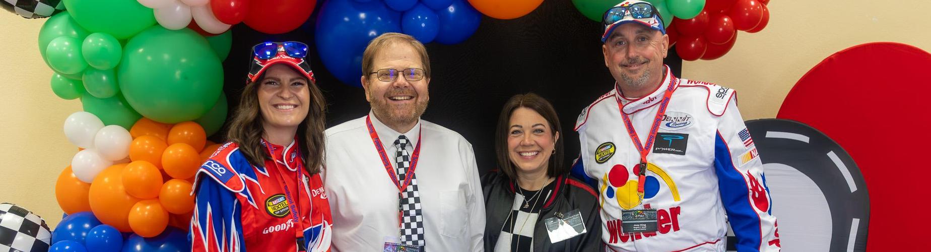 Four people in racing outfits pose in front of colorful balloons and a road-themed backdrop.