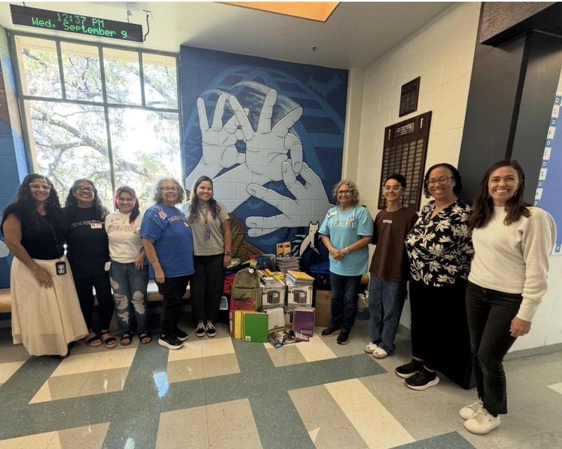 4 HS staff, 4 TDF members, and 1 HS student smiling next to a pile of school supplies in between of the crowd