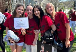 Four female students in red shirts pose outdoors, holding a certificate and a painting.