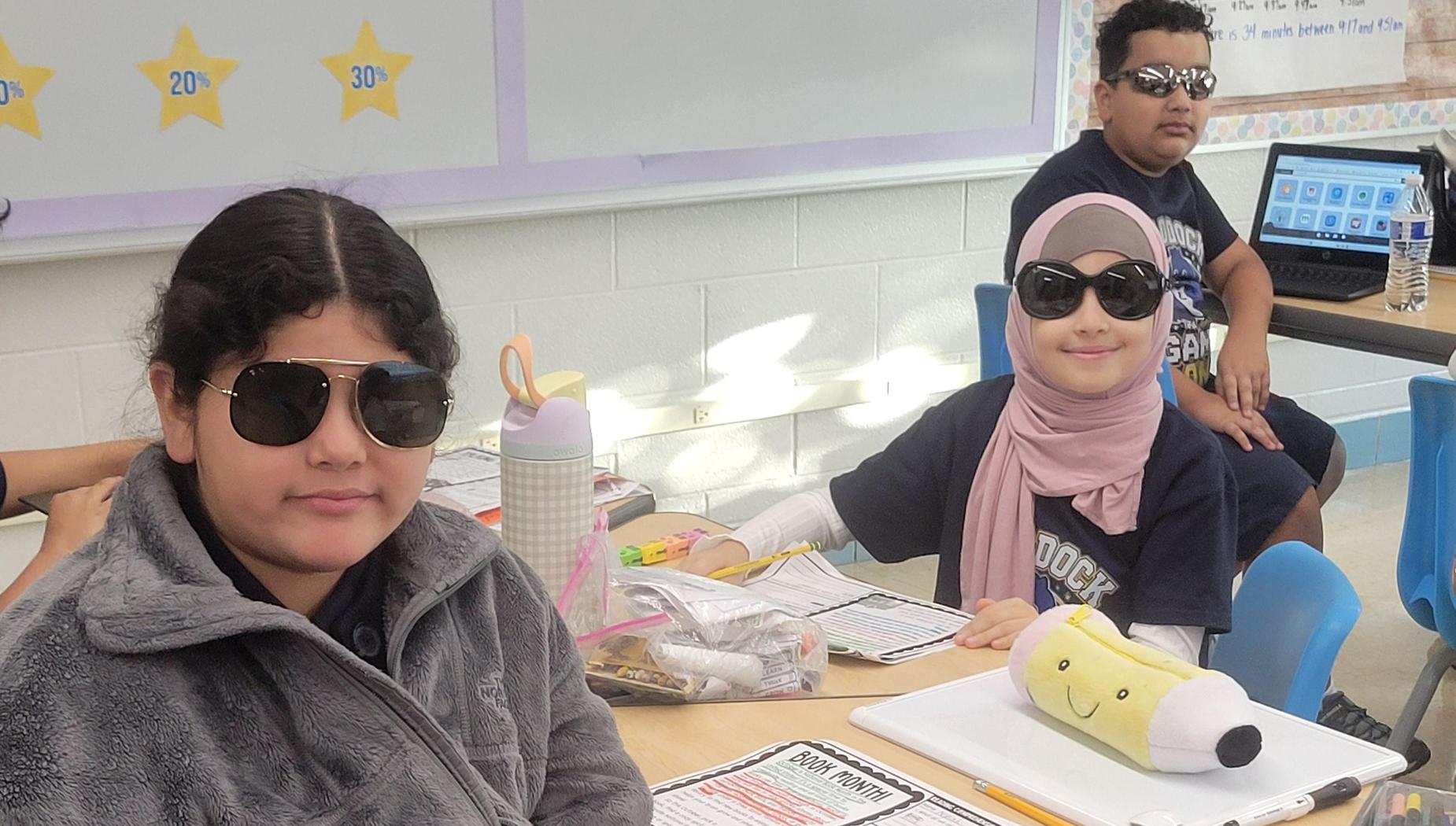 Two students at a classroom table with worksheets and colorful supplies.