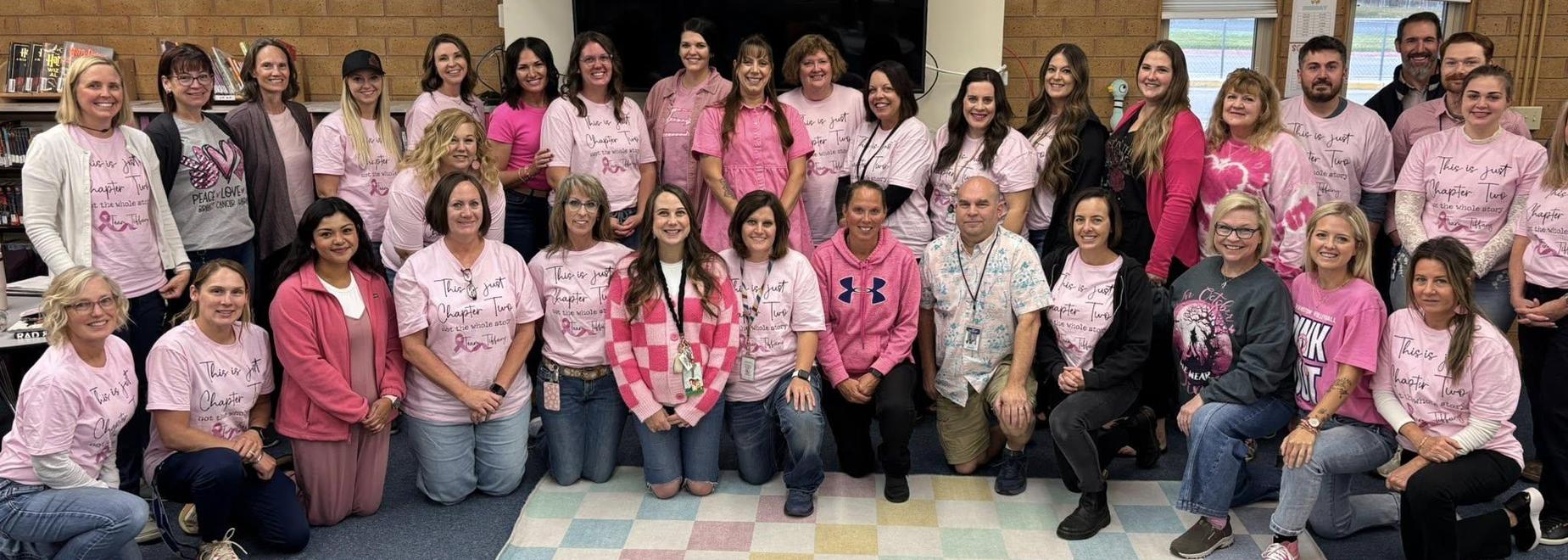 Group of people wearing pink shirts posing together for a photo in a classroom setting.