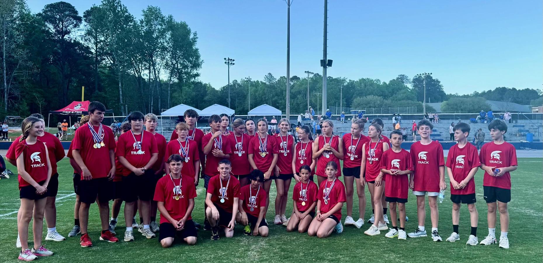 Team of young athletes in red shirts, displaying medals at a track event.