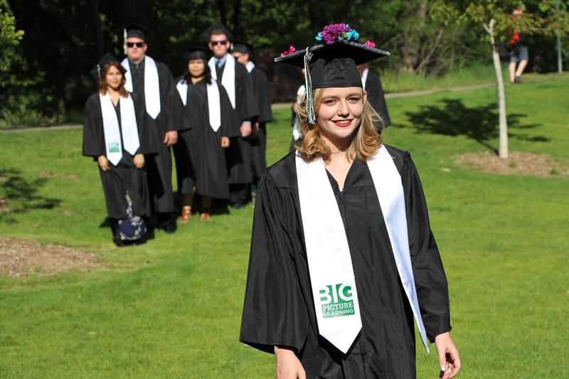 A smiling high school graduate in black cap and gown, with a white cowl that reads Big Picture Learning, walks through a park on a sunny day