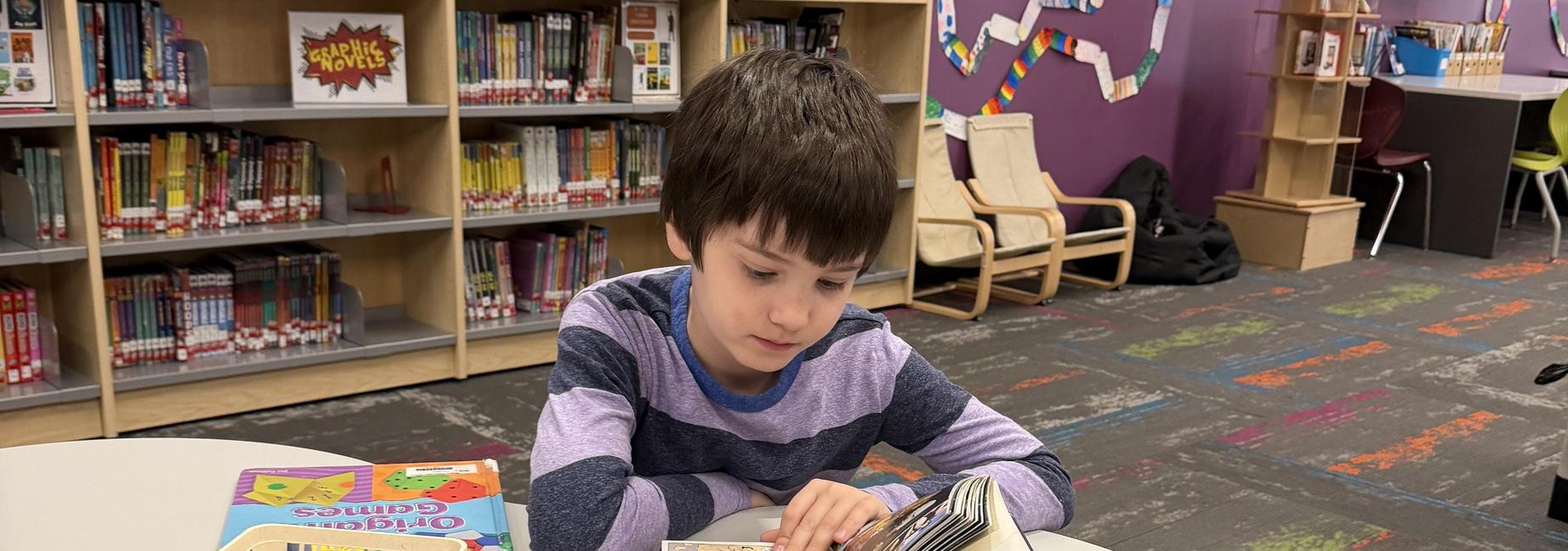 A young boy reading a colorful comic book at a table in a library.
