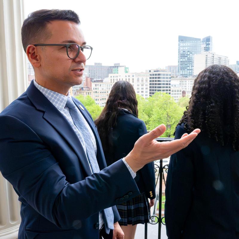 A man in a suit gestures while speaking, with two students in uniforms behind him.
