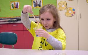 A girl in a yellow shirt stirs a mixture in a cup with a stick, focusing intently.