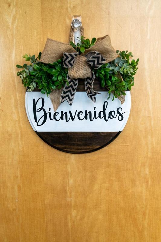 Circular wooden sign with a white band that reads “Bienvenidos” with a bow and greenery over it hangs on a door at Arco Iris charter school.