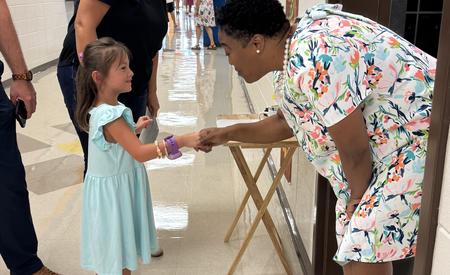 A smiling woman greets a young girl in a light blue dress in a school hallway.