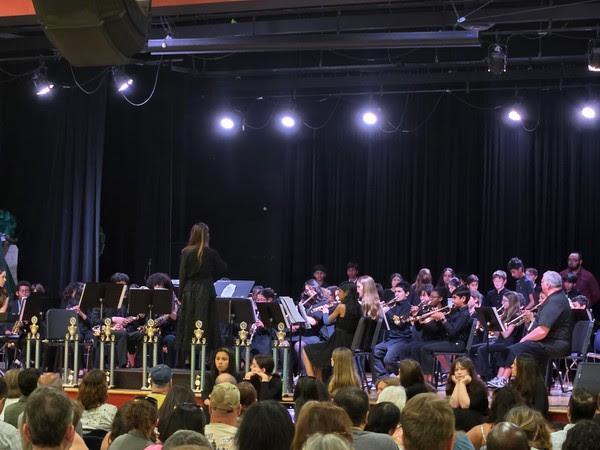 A conductor stands in front of students, all in black, playing instruments in band.