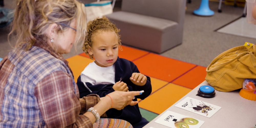 An adult and a child interact while sitting on the floor, focusing on cards and a marker in a classroom.