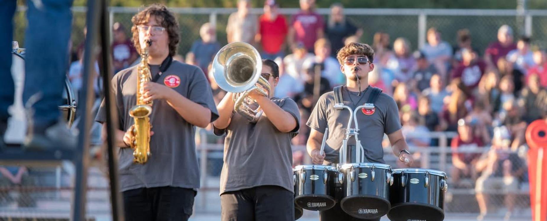 Three musicians playing brass and percussion instruments on a field at a crowded event.