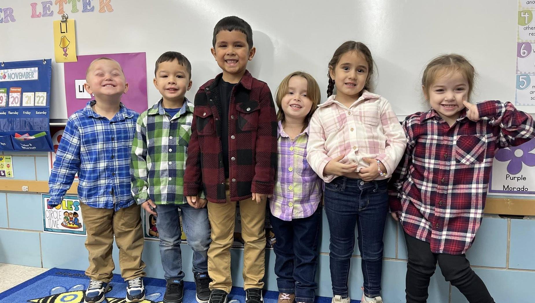 Six children in plaid shirts posing together in a classroom