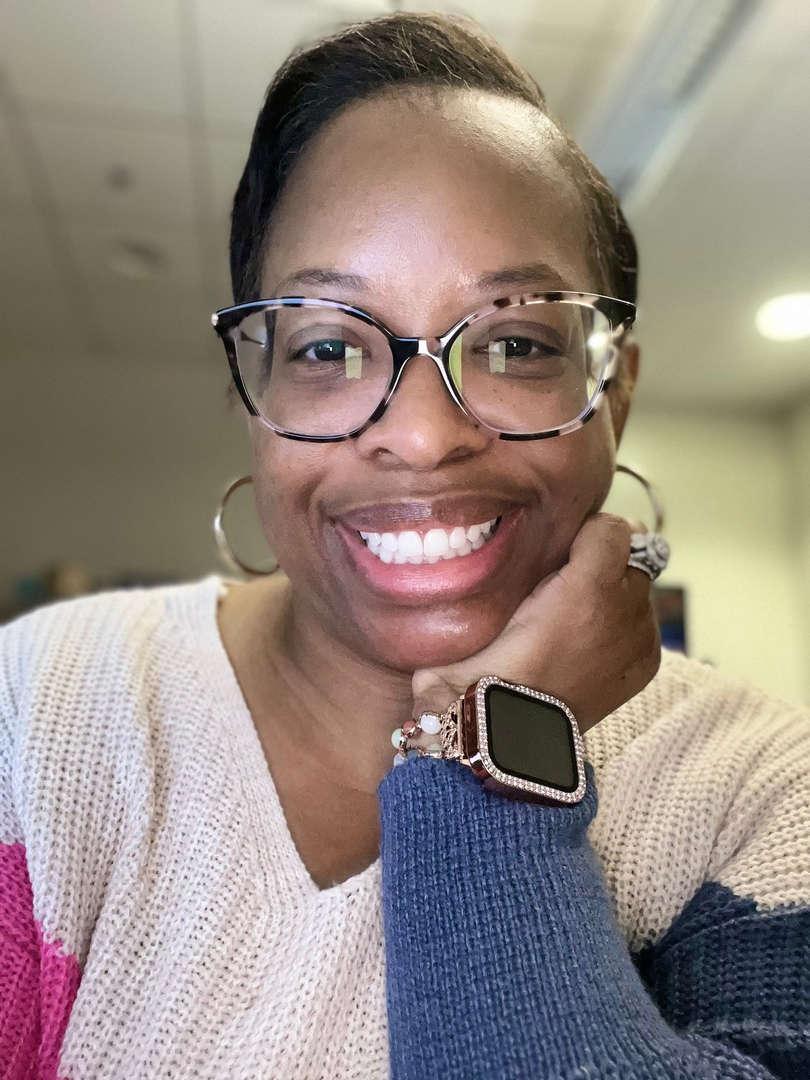 A smiling woman with glasses, resting her chin on her hand in a cozy sweater.