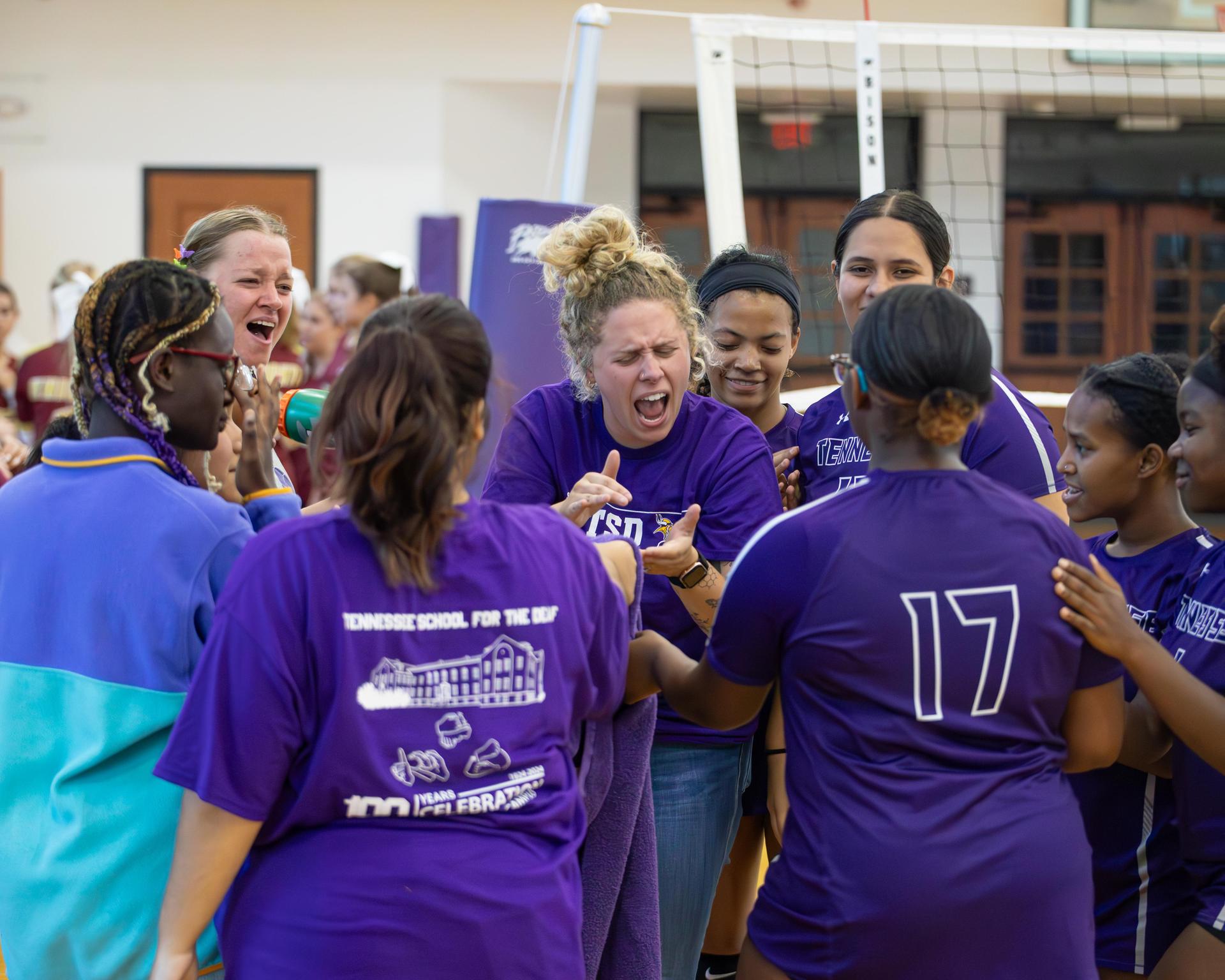 Cheering players in purple jerseys celebrate a team's victory in a gymnasium.