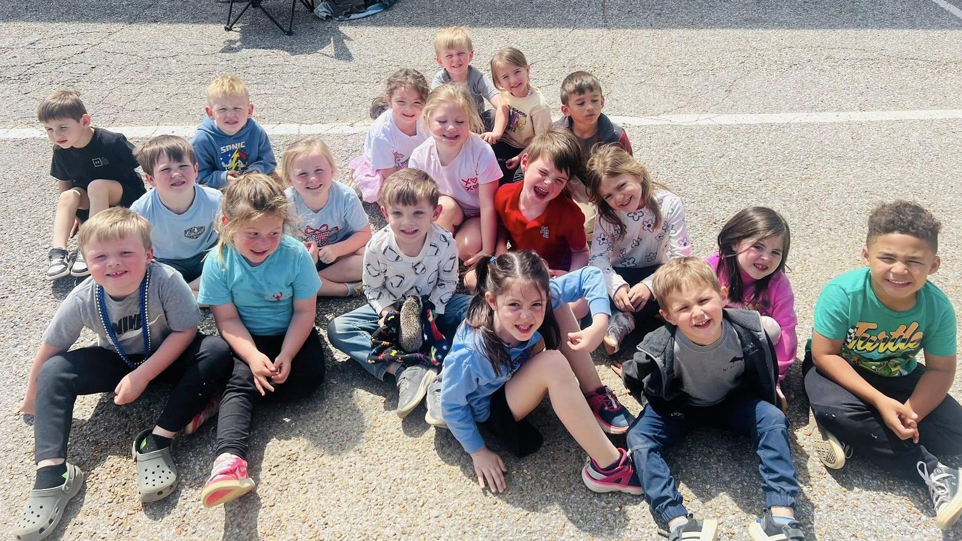 A large group of children sits together and smiles on a sunny day.
