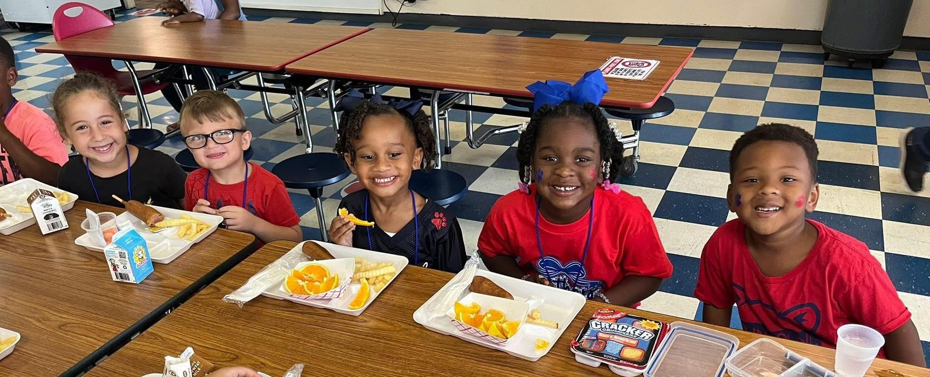 Five smiling children in red shirts at a lunch table enjoying their food.