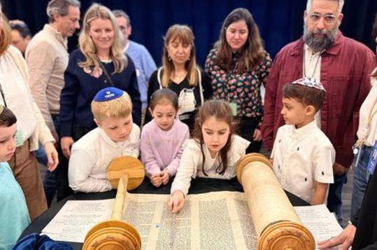 Children and adults gathered around a Torah scroll in a celebratory setting.