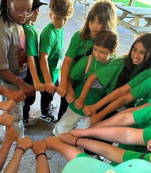 A diverse group of children and adults joining fists together in a circle, wearing green shirts.