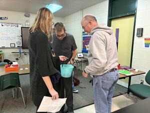 Three students gather around a bucket in a classroom while discussing.