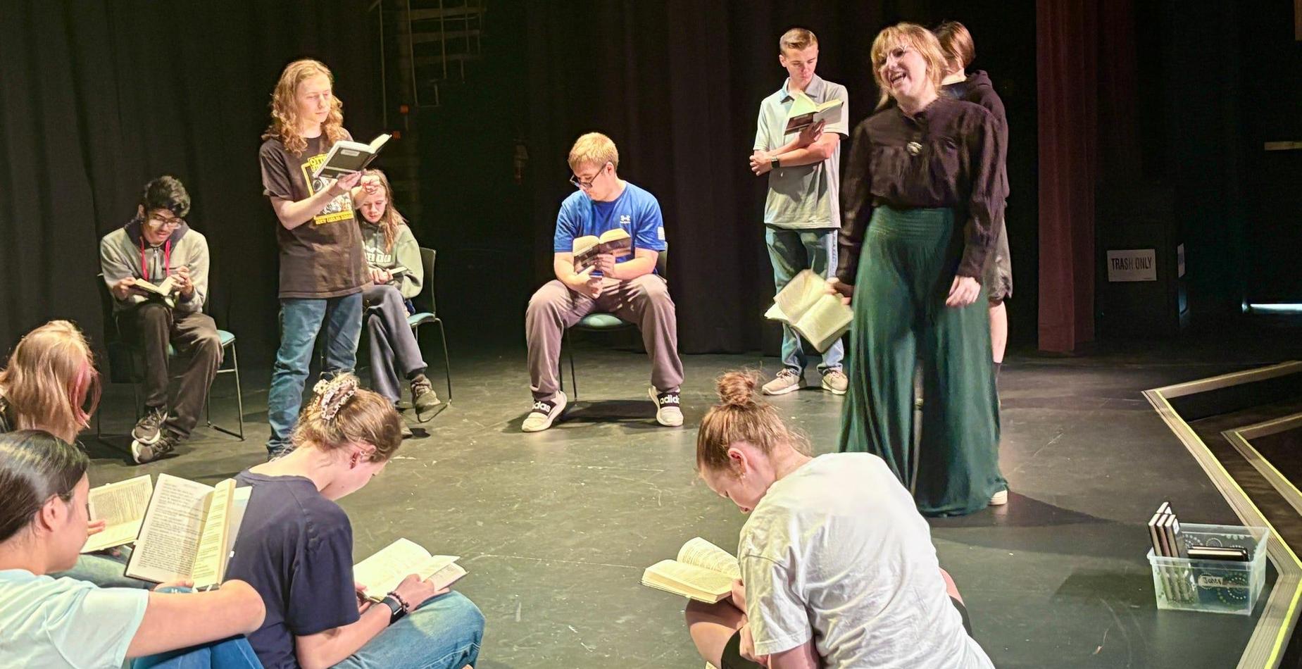 A group of young actors rehearsing a scene on stage with script books.