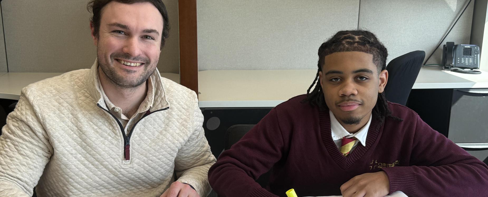Two men sitting at a table, one smiling and the other highlighting a document.
