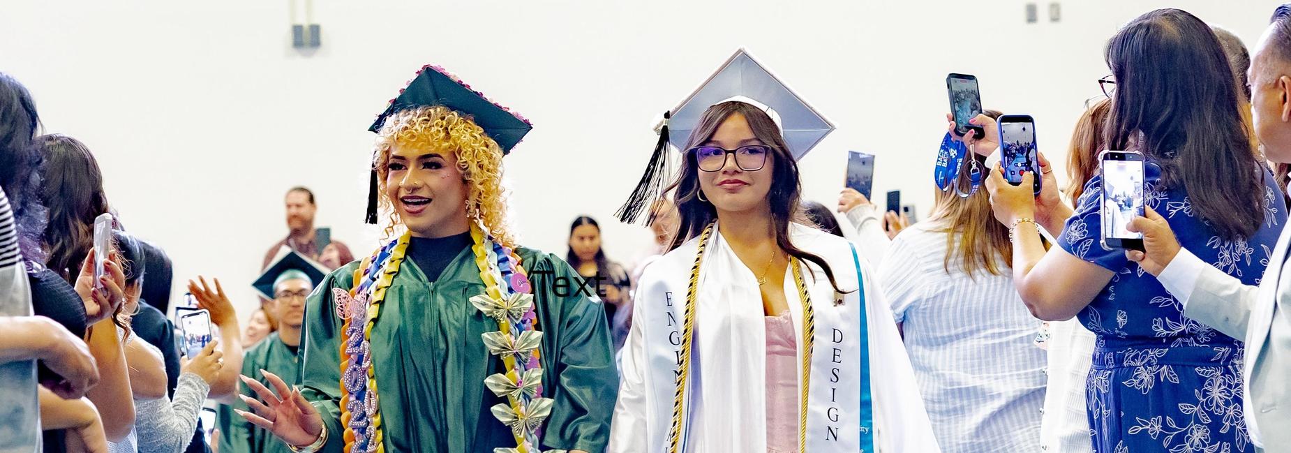 Two graduates walk side by side down a gymnasium aisle during a commencement processional. The graduate on the left wears a green gown with an elaborate braided and butterfly-adorned lei. The one on the right wears a white gown with a sash reading "Engineering Design" and a gold honor cord, representing the Eastside High School STEM Magnet Class of 2026. Family members line both sides holding up phones to capture the moment.