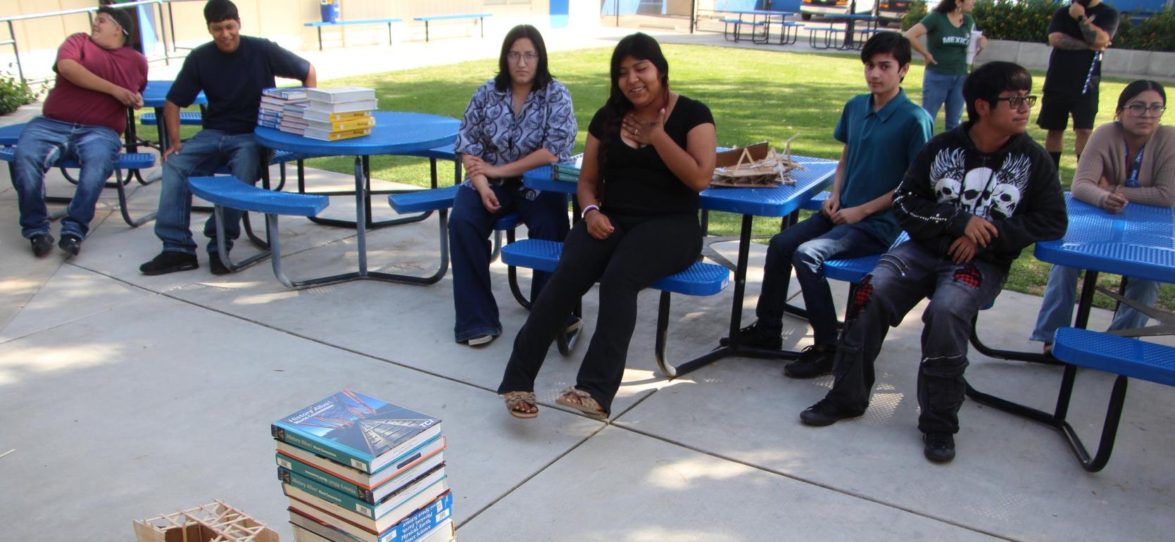 A tower of stacked books on a concrete surface with people seated nearby.