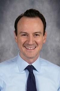 Smiling man in a white shirt and patterned tie against a gray background.